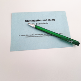 Ein blauer Briefwahlumschlag auf dem ein grüner Kugelschreiber mit dem Logo der Stadt Erftstadt liegt