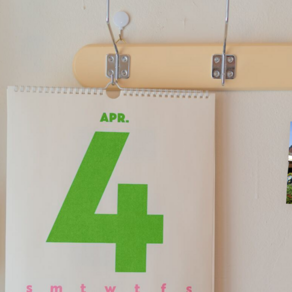 Ein Wandkalender mit einer großen grünen 4 hängt an einer Vintage-Garderobe mit drei Haken. Daneben eine Postkarte an der Wand mit einer Marktszene aus Lechenich