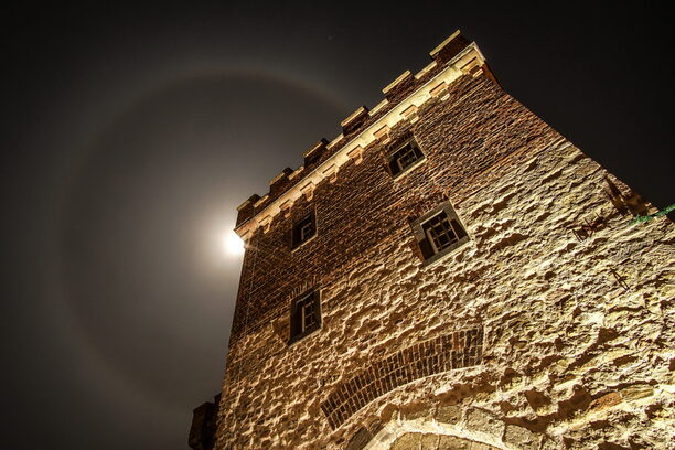 Nachtaufnahme von beleuchtetem Stadttor, extreme Untersicht, hinter der Zinnen leuchtet der Mond hervor, der von einer großen Korona umgeben ister eine runde