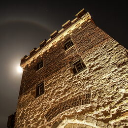 Nachtaufnahme von beleuchtetem Stadttor, extreme Untersicht, hinter der Zinnen leuchtet der Mond hervor, der von einer großen Korona umgeben ister eine runde