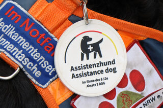 Runde Plakette mit der Inschrift "Assistenzhund im Sinne des § 12a Absatz 3 BGG". Darüber ist ein schwarzer Hund mit weißem Kreuz auf dem Laib abgebildet. Über diesem spannt sich ein dünner Bogen in den deutschen Nationalfarben.