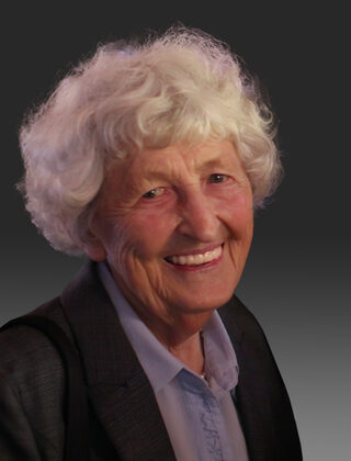 Porträt einer freundlich lächelnden alten Dame mit weißem, gelockten Haar.