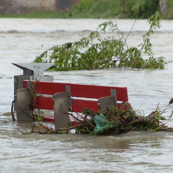 Eine rote Sitzbank ist bis zur Hälfte überschwemmt, Äste und Müll wurden an die Bank gespült und hängen dort fest. Ein Baum liegt im Wasser