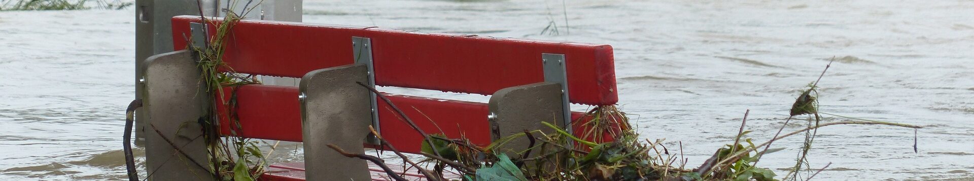 Eine rote Sitzbank ist bis zur Hälfte überschwemmt, Äste und Müll wurden an die Bank gespült und hängen dort fest. Ein Baum liegt im Wasser