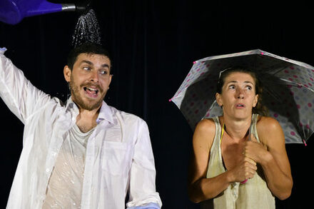 Symbolbild des 12 Stufen Theaters: Ein Mann schüttet sich mit einer blauen Gießkanne Wasser über den Kopf. Eine Frau steht daneben und schützt sich mit einem Regenschirm.